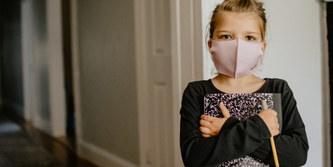 A child wearing a mask holds a notebook and a pencil